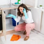 A women in front of her kitchen sink is calling a plumber on her cell phone because she's having a holiday plumbing emergency where there is a leak in the pipe