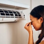 A woman pinching her nose in front a smelly home air conditioner vent.